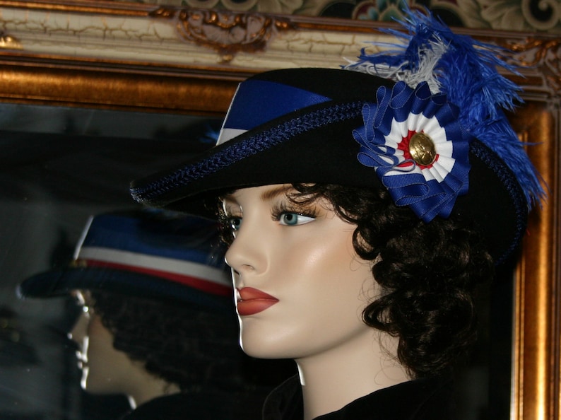 May include: A black hat with blue ribbon and feather detailing. The hat has a blue, white and red rosette with a gold button. The hat is displayed on a mannequin.