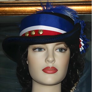 May include: A black hat with a wide brim, decorated with a blue and white striped ribbon, red ribbon, and gold buttons. The hat has a blue feather and a decorative ribbon on the side. The hat is worn by a mannequin head.