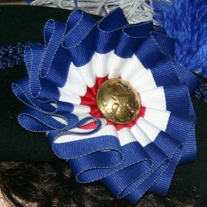 May include: A decorative rosette made of blue, white, and red striped ribbon, with a gold-coloured button at the centre. The rosette is attached to a black surface, possibly a hat. The ribbon is arranged in a circular pattern.