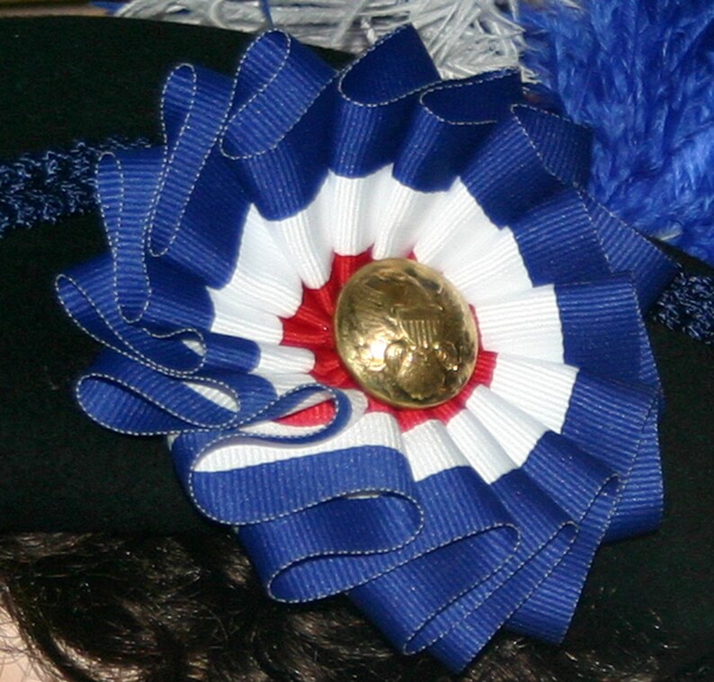 May include: A decorative rosette made of blue, white, and red striped ribbon, with a gold-coloured button at the centre. The rosette is attached to a black surface, possibly a hat. The ribbon is arranged in a circular pattern.