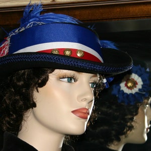 May include: A decorative hat in blue, white, and red, adorned with gold buttons and feathers. The hat features a black brim and a blue band. The hat is displayed on a mannequin head. A mirror reflects the hat's image.