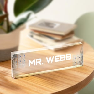 Könnte beinhalten: Ein klares Acryl-Namensschild für den Schreibtisch mit dem Text "MR. WEBB" in weißen Buchstaben. Der Hintergrund des Namensschilds hat ein dezentes Muster aus blauem und weißem Binärcode.
