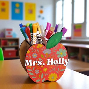 Puede incluir: Un organizador de escritorio de madera con forma de manzana, con un diseño floral rojo y el nombre "Mrs. Holly" en blanco. Contiene varios rotuladores y bolígrafos de colores. El fondo muestra un aula.