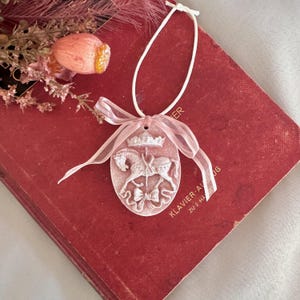 Peut inclure: Un ornement suspendu ovale rose pâle présentant un motif en relief d'un cheval de carrousel surmonté d'une couronne et orné d'un nœud. L'ornement est suspendu par un cordon blanc et décoré d'un ruban rose clair transparent noué en boucle.