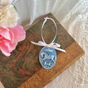 Puede incluir: Adorno ovalado azul claro con un diseño de caballo y corona, atado con una cinta blanca. El adorno descansa sobre un libro antiguo con una cubierta veteada, junto a flores rosas y blancas.