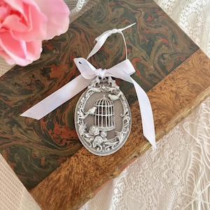 Peut inclure: Ornement ovale argenté avec un motif de cage à oiseaux, deux oiseaux et des détails floraux. Il est attaché avec un ruban blanc et posé sur un livre ancien à couverture marbrée.