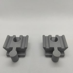 Può includere: Due pezzi grigi di binari di treni giocattolo in plastica sono mostrati su uno sfondo bianco. Ogni pezzo di binario ha una base rettangolare con un perno circolare che si estende verso il basso e un foro circolare nella parte superiore.