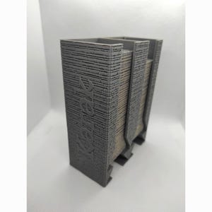 May include: A grey, rectangular pencil holder with a textured brick design. The word "Yarek" is embossed on the side. The holder has a unique architectural style, with a light brown accent on the side.