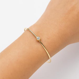 Könnte beinhalten: Ein zartes goldfarbenes Armband mit einem einzelnen, runden, klaren Edelstein. Das dünne, gebogene Band des Armbands ist goldfarben. Der Edelstein ist in einer kleinen, kreisförmigen Goldfassung gefasst. Das Armband wird am Handgelenk getragen.