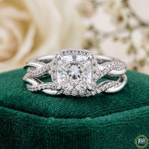 Peut inclure: Une bague de fiançailles argentée avec une pierre centrale taille coussin entourée d'un halo de petites pierres rondes. L'anneau présente un design torsadé incrusté de pierres pavées, accompagné d'une alliance assortie profilée.
