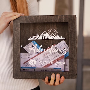 May include: A dark wooden shadow box displaying travel tickets. The box has the text "The Adventures Of Kate & Jason" with a mountain and sun graphic. The tickets are visible through a clear front, a keepsake for memories.