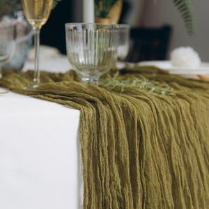 May include: A green linen table runner with a textured, crinkled look. The runner is draped over a white tablecloth and is perfect for adding a touch of rustic elegance to any table setting.