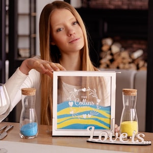 May include: A white wooden sand ceremony box with a glass front. The box has a white inscription that reads "The Collins Family" with a heart design. Two glass bottles with cork stoppers are on either side of the box. One bottle contains blue sand and the other contains yellow sand. A wooden sign with the text "Love is..." is in front of the box.