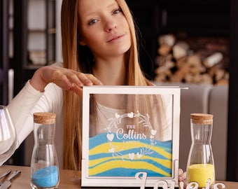 Könnte beinhalten: Eine weiße Sandzeremonie-Box aus Holz mit einer Glasfront. Die Box trägt eine weiße Inschrift mit dem Text "The Collins Family" und einem Herzdesign. Zwei Glasflaschen mit Korkstopfen befinden sich auf beiden Seiten der Box. Eine Flasche enthält blauen Sand und die andere gelben Sand. Ein Holzschild mit dem Text "Love is..." steht vor der Box.