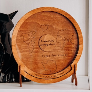 May include: A circular wooden coin holder with a world map design. The holder has a light brown finish and features the phrases "A coin from every place", "Time for travel", and "Travel far, collect more". It is supported by a wooden stand.