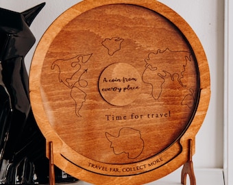 May include: A round, wooden travel coin holder with a world map design. The holder has a light brown finish and includes the phrases "A coin from every place", "Time for travel", and "Travel far, collect more". It is supported by a wooden stand.