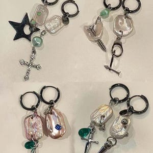 Può includere: Quattro paia di orecchini unici con cerchi neri. Ogni paio presenta perle bianche irregolari, perline verdi e vari ciondoli, tra cui una stella, una croce, una vite e un pugnale. Gli orecchini hanno un'estetica moderna e audace.