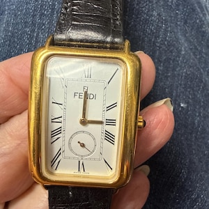 May include: A rectangular gold-tone wristwatch featuring a white dial with black Roman numeral hour markers, a sub-dial at the bottom, and the Fendi logo at the top. The watch includes a black textured leather strap and a small crown on the right side.