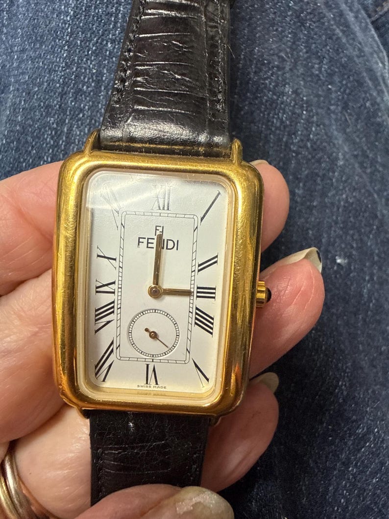 May include: A rectangular gold-tone wristwatch featuring a white dial with black Roman numeral hour markers, a sub-dial at the bottom, and the Fendi logo at the top. The watch includes a black textured leather strap and a small crown on the right side.
