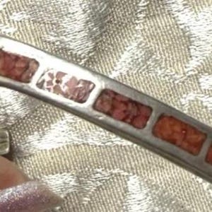 May include: Silver-tone cuff bracelet with turquoise and red-orange inlaid stones. The bracelet has a V-shape design with rectangular stones set in a silver metal frame. The stones are arranged in a repeating pattern.