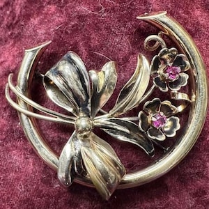 May include: A gold-toned brooch with a horseshoe shape, featuring a stylized bow and two flowers with pink centers. The brooch has a vintage aesthetic and is set against a burgundy background.