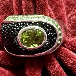 May include: A silver ring with a central oval green gemstone, surrounded by black stones and green accents. The ring is set against a red velvet background, showcasing its intricate design and color contrast.