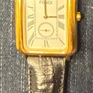 May include: A rectangular gold-tone wristwatch with a white face featuring Roman numeral markers and the Fendi logo. The watch has a textured dark brown leather strap with a gold-tone buckle engraved with the Fendi brand name at the top.