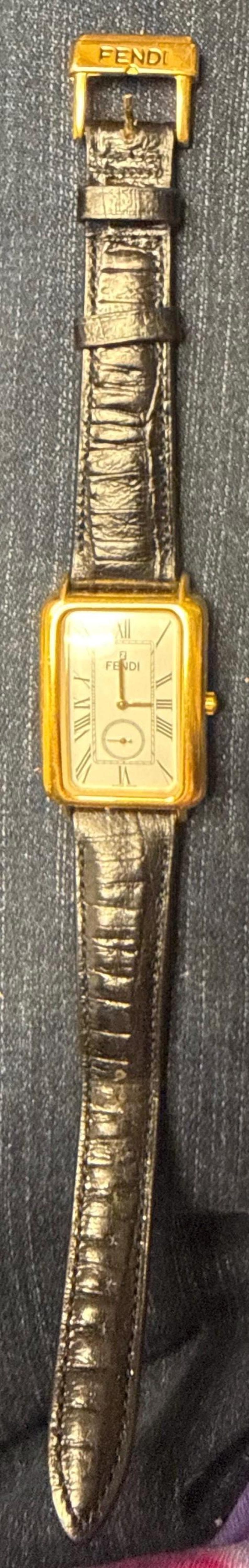 May include: A rectangular gold-tone wristwatch with a white face featuring Roman numeral markers and the Fendi logo. The watch has a textured dark brown leather strap with a gold-tone buckle engraved with the Fendi brand name at the top.