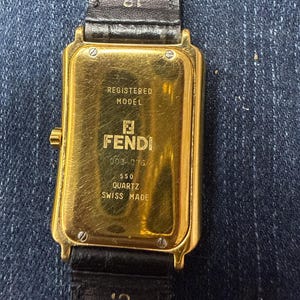 May include: A rectangular gold-tone Fendi quartz watch with a black leather strap. The case back features the text REGISTERED MODEL, FENDI, 0034076, 550, QUARTZ, and SWISS MADE. The strap is marked with the number 18 near the watch case lugs.