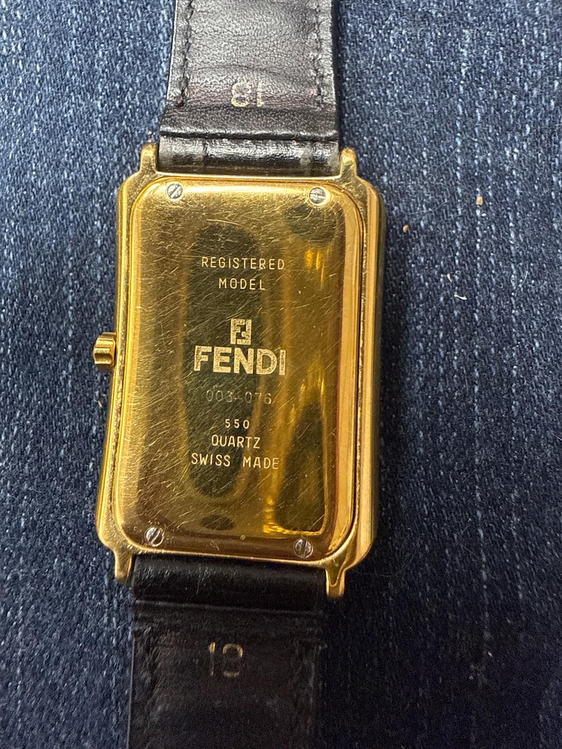 May include: A rectangular gold-tone Fendi quartz watch with a black leather strap. The case back features the text REGISTERED MODEL, FENDI, 0034076, 550, QUARTZ, and SWISS MADE. The strap is marked with the number 18 near the watch case lugs.