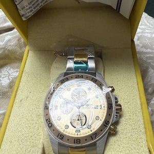 May include: A silver and gold Invicta chronograph watch with a gold dial, featuring multiple sub-dials and a date display. The watch is in its original yellow box. The watch band has silver, gold, and teal accents.
