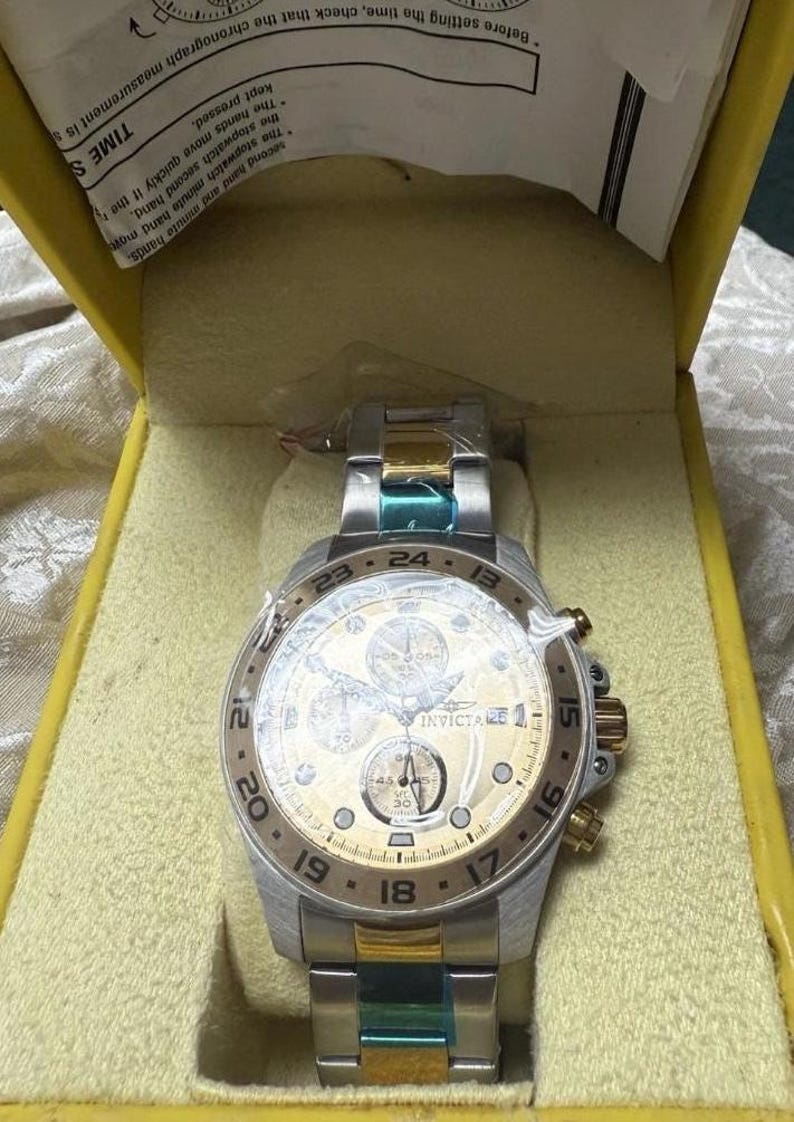 May include: A silver and gold Invicta chronograph watch with a gold dial, featuring multiple sub-dials and a date display. The watch is in its original yellow box. The watch band has silver, gold, and teal accents.