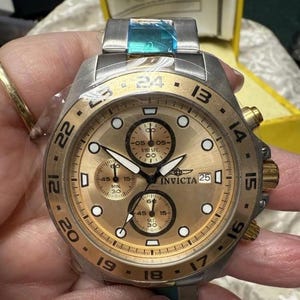 May include: A gold-toned Invicta chronograph watch with a stainless steel band. The watch face features multiple sub-dials, luminous markers, and the Invicta logo. The bezel has black numerals and gold accents. The watch is new, with protective film.