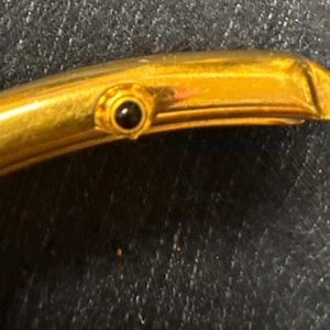 May include: A gold-toned watch case with a curved, rectangular profile. The side features a small, circular winding crown set with a dark, round stone accent. The metal surface has a polished finish, and the watch is attached to a dark strap.