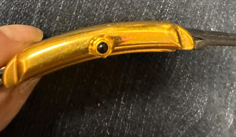 May include: A gold-toned watch case with a curved, rectangular profile. The side features a small, circular winding crown set with a dark, round stone accent. The metal surface has a polished finish, and the watch is attached to a dark strap.