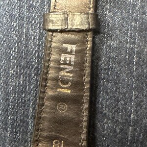 May include: A black leather watch strap with a gold-tone metal buckle featuring the FENDI logo. The strap is embossed with FENDI, a registered trademark symbol, and the number 18. The watch case back is gold-tone metal with REGISTERED MODEL and FENDI.