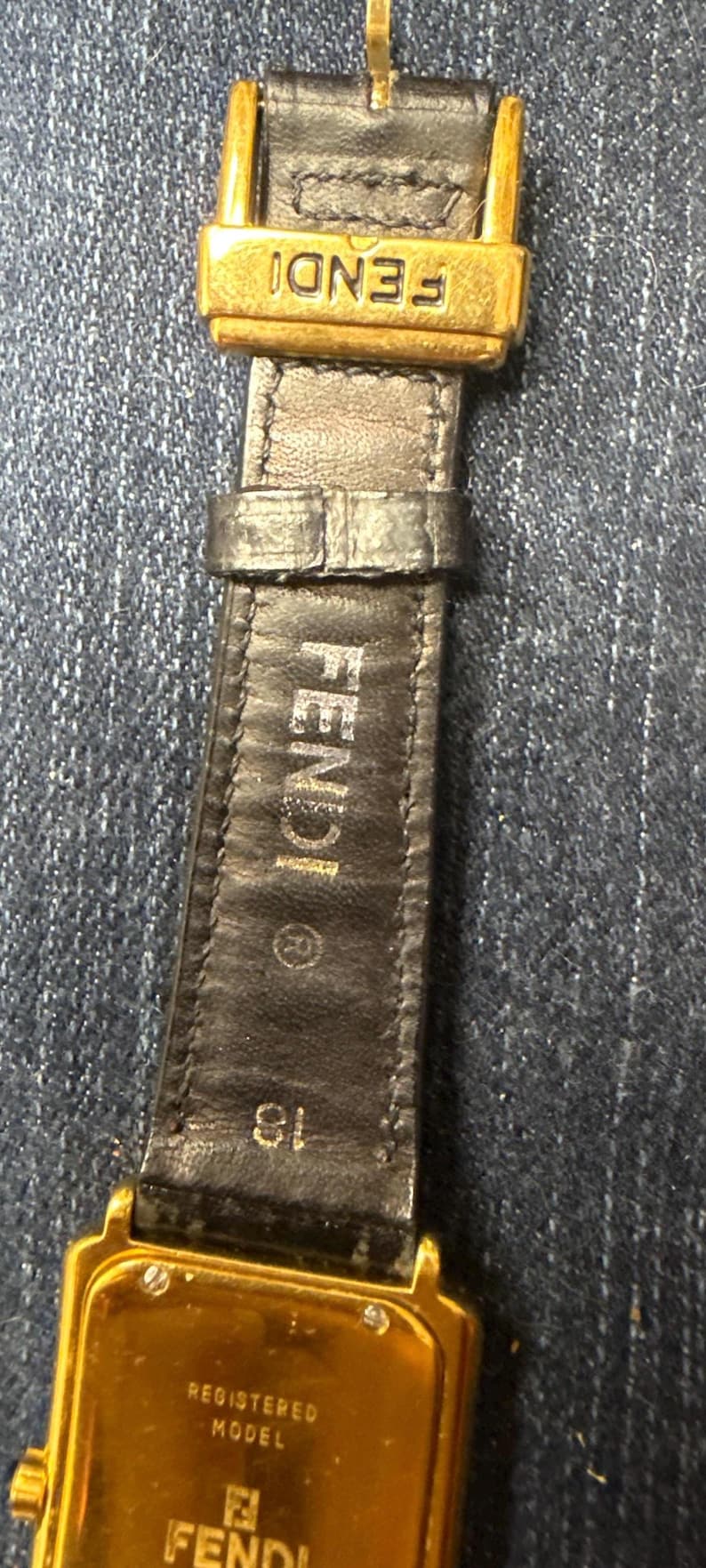 May include: A black leather watch strap with a gold-tone metal buckle featuring the FENDI logo. The strap is embossed with FENDI, a registered trademark symbol, and the number 18. The watch case back is gold-tone metal with REGISTERED MODEL and FENDI.