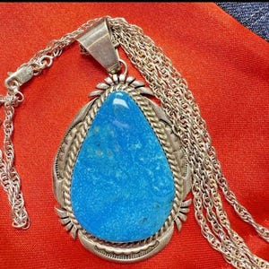 May include: A silver pendant necklace featuring a large, teardrop-shaped turquoise stone. The stone is set in a detailed silver frame with a braided edge and is suspended from a delicate silver chain. The necklace is displayed on a red surface.