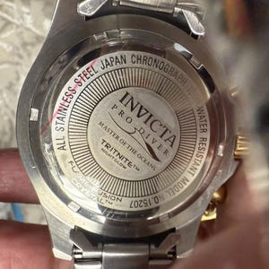 May include: Close-up of a stainless steel Invicta Pro Diver watch. The watch features a silver band and a round face with the text "All Stainless Steel Japan Chronograph 100M Water Resistant". The watch also has the text "Master of the Oceans" and "Tritnite Nightglow".
