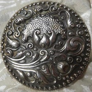 May include: A silver-toned, round, decorative brooch with an intricate floral design. The design features a central flower with a textured center and swirling leaves.