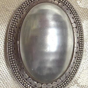 May include: Oval-shaped silver brooch with a large, lustrous, pearly-gray center stone. The brooch has a detailed border with small, round beads and a decorative edge. The design is symmetrical and elegant, suitable for formal wear.