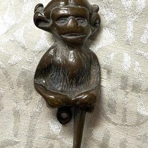 May include: Bronze-toned door knocker in the shape of a whimsical goblin-like figure. The figure has a detailed face with large eyes, curly hair, and is seated with its arms crossed. A ring is at the top for hanging.