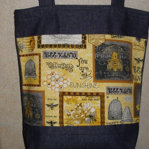 May include: A blue denim tote bag with a yellow and black bee-themed fabric lining. The fabric features a honeycomb pattern, bees, and the text "Bee Kind", "Sweet to the Soul", "You are my Sunshine", "The Life of a Honey Bee", and "The Flowers and the Bees".