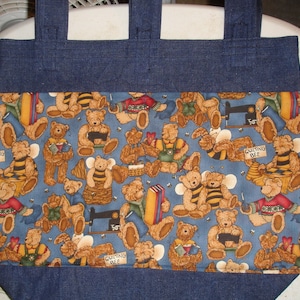 May include: A blue denim tote bag with a fabric lining featuring a repeating pattern of bears and bees. The fabric lining is blue with a white background. The bears are wearing different colored clothing and holding various items, including books, sewing machines, and baskets. The text "Quilting Bee" is printed on two signs in the pattern.
