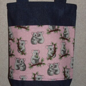 May include: A denim tote bag with a pink fabric lining featuring a repeating pattern of gray koalas hugging on tree branches. The koalas are on a pink background with green leaves and white hearts.