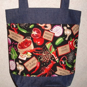 May include: A blue denim tote bag with a black fabric lining featuring a colorful print of various Cajun and Creole food ingredients, including shrimp, crawfish, tomatoes, onions, peppers, and spices. The fabric lining also includes text labels such as "Shrimp Bisque", "Hot 'N Spicy Gumbo", "Jambalaya", and "Cajun Creole".