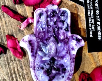 May include: Purple and white hamsa hand charm with a floral design. The hamsa hand is a symbol of protection and good luck.