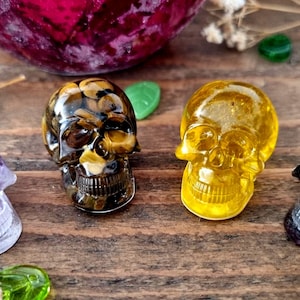May include: Four crystal skull figurines in different colours: purple, brown, yellow, and black. The skulls are carved from natural stones and have intricate details.