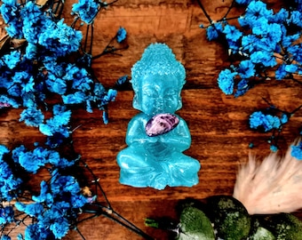 May include: A teal blue resin Buddha statue holding a purple and white crystal in its hands. The statue is sitting on a wooden surface.