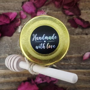 以下が含まれることがあります： 金色の蓋と、白い筆記体で「Handmade with love」と書かれた黒いラベルが付いた瓶。木製のハニーディッパーが瓶の横に置かれています。乾燥したバラの花びらが木製の表面に散らばっています。
